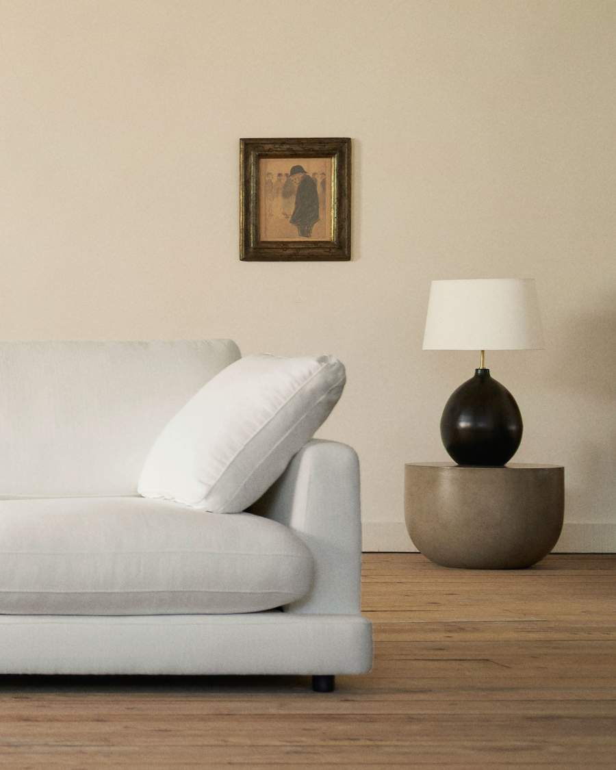 Gros plan de l'accoudoir et d'un coussin du canapé Gala blanc, avec une lampe et une table ronde.