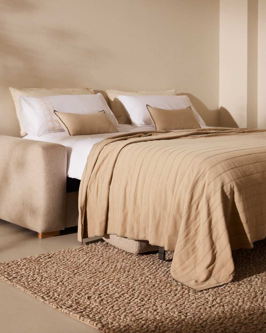 Canapé Anley déplié en lit avec draps blancs, couverture beige et coussins, vu de côté.