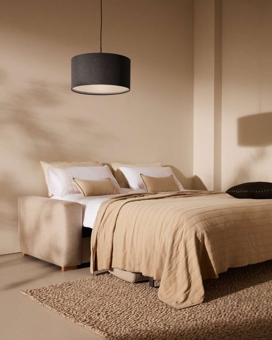 Canapé Anley déplié en lit double avec draps blancs, couverture beige et coussins, dans une chambre.