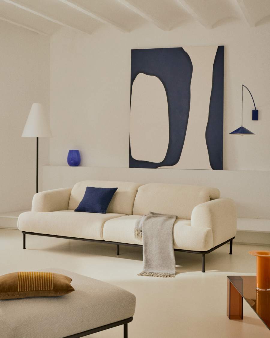 Canapé Aurum crème aux pieds noirs dans un salon blanc, avec coussin bleu foncé et tableau abstrait.