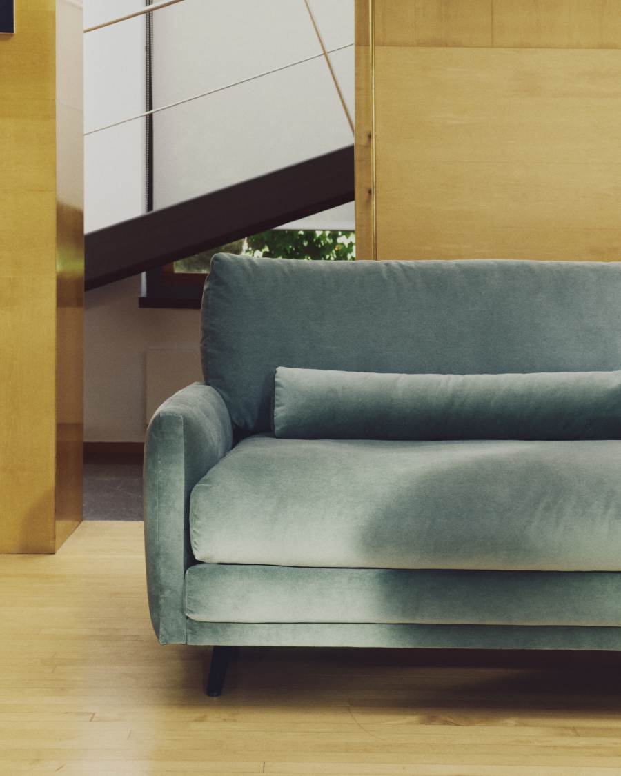 Gros plan de l'accoudoir arrondi et de l'assise du canapé en velours bleu-gris dans une pièce en bois.