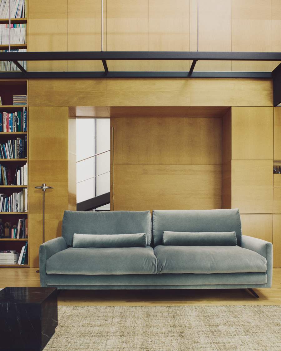 Canapé Veliro en velours bleu-gris devant une bibliothèque et des murs en bois clair.