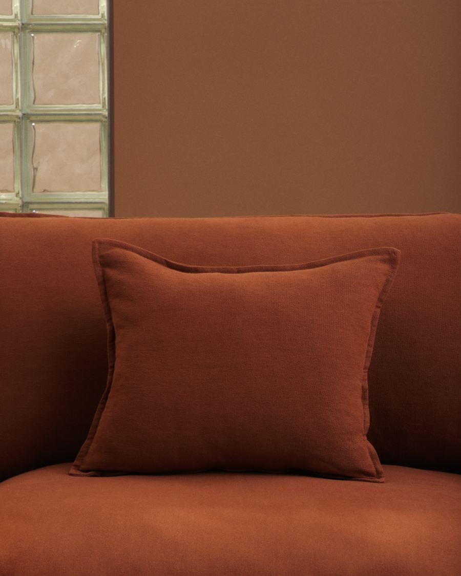 Gros plan d'un coussin terracotta posé sur le dossier du canapé Anarela, près d'une fenêtre en verre.